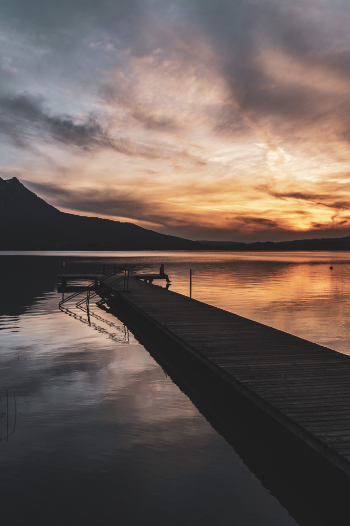 Am Steg beim Badeplatz Loibichl am Mondsee mit Sonnenuntergang und Drachenwand im Hintergrund an der Grenze von Salzburg und Oberösterreich, Österreich.