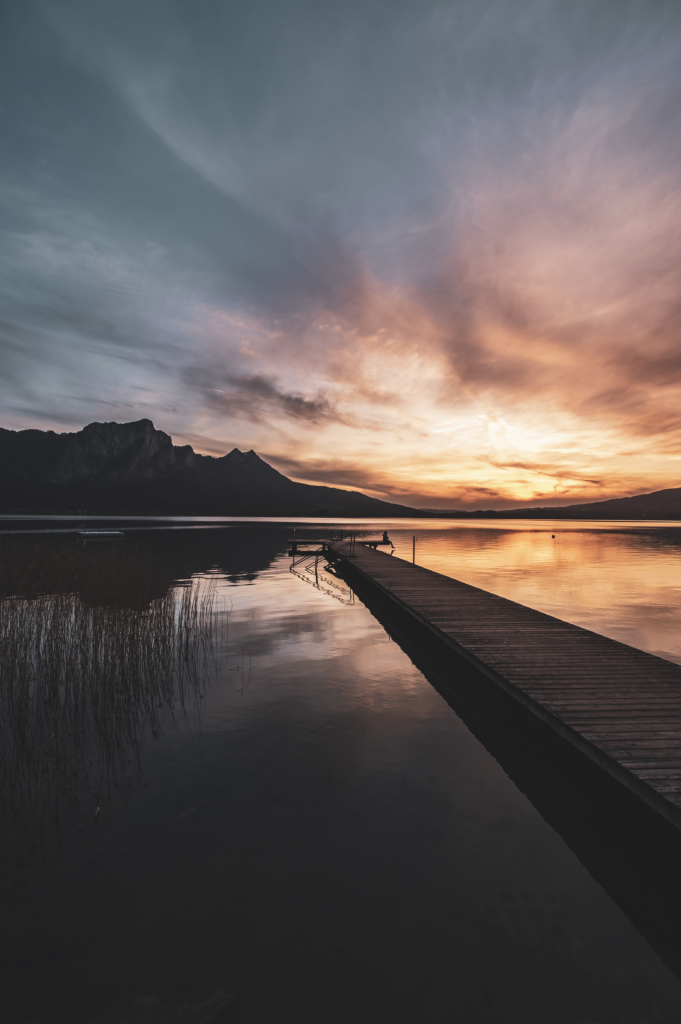 Am Steg beim Badeplatz Loibichl am Mondsee mit Sonnenuntergang und Drachenwand im Hintergrund an der Grenze von Salzburg und Oberösterreich, Österreich.