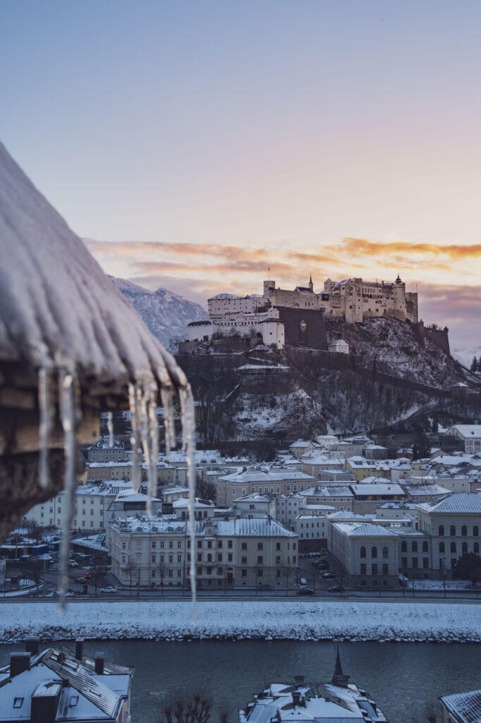 Blick vom Kapuzinerberg auf die verschneite Stadt Salzburg und die Festung Hohensalzburg während eines Sonnenuntergang im Winter.