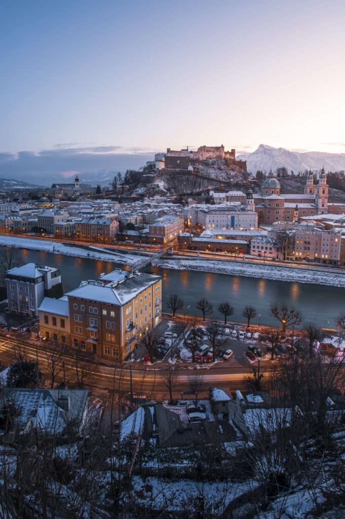 Blick vom Kapuzinerberg auf die verschneite Stadt Salzburg und die Festung Hohensalzburg während eines Sonnenuntergang im Winter.