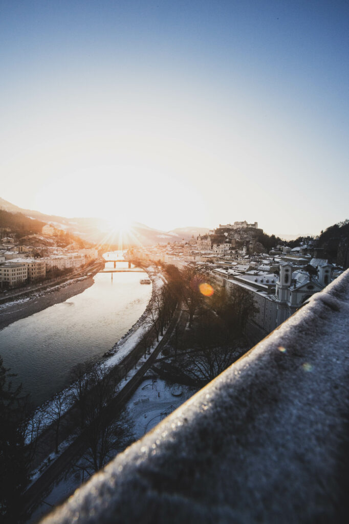 Blick vom Mönchsberg auf die verschneite Stadt Salzburg und die Festung Hohensalzburg während eines Sonnenaufgangs im Winter.