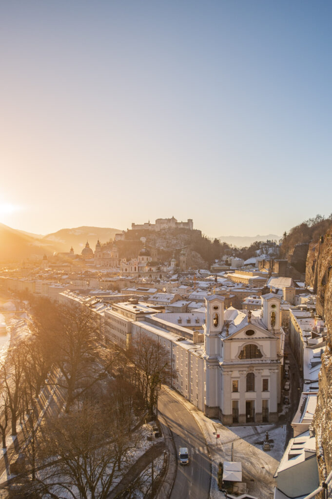 Blick vom Mönchsberg auf die verschneite Stadt Salzburg und die Festung Hohensalzburg während eines Sonnenaufgangs im Winter.