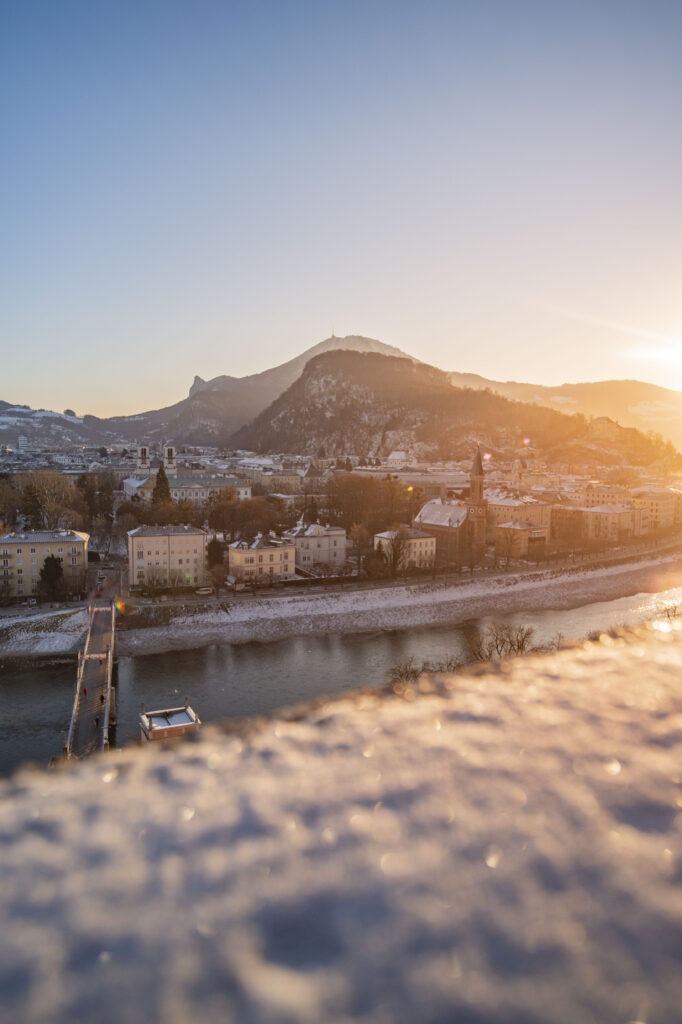 Blick vom Mönchsberg auf die verschneite Stadt Salzburg und den Gaisberg während eines Sonnenaufgangs im Winter.