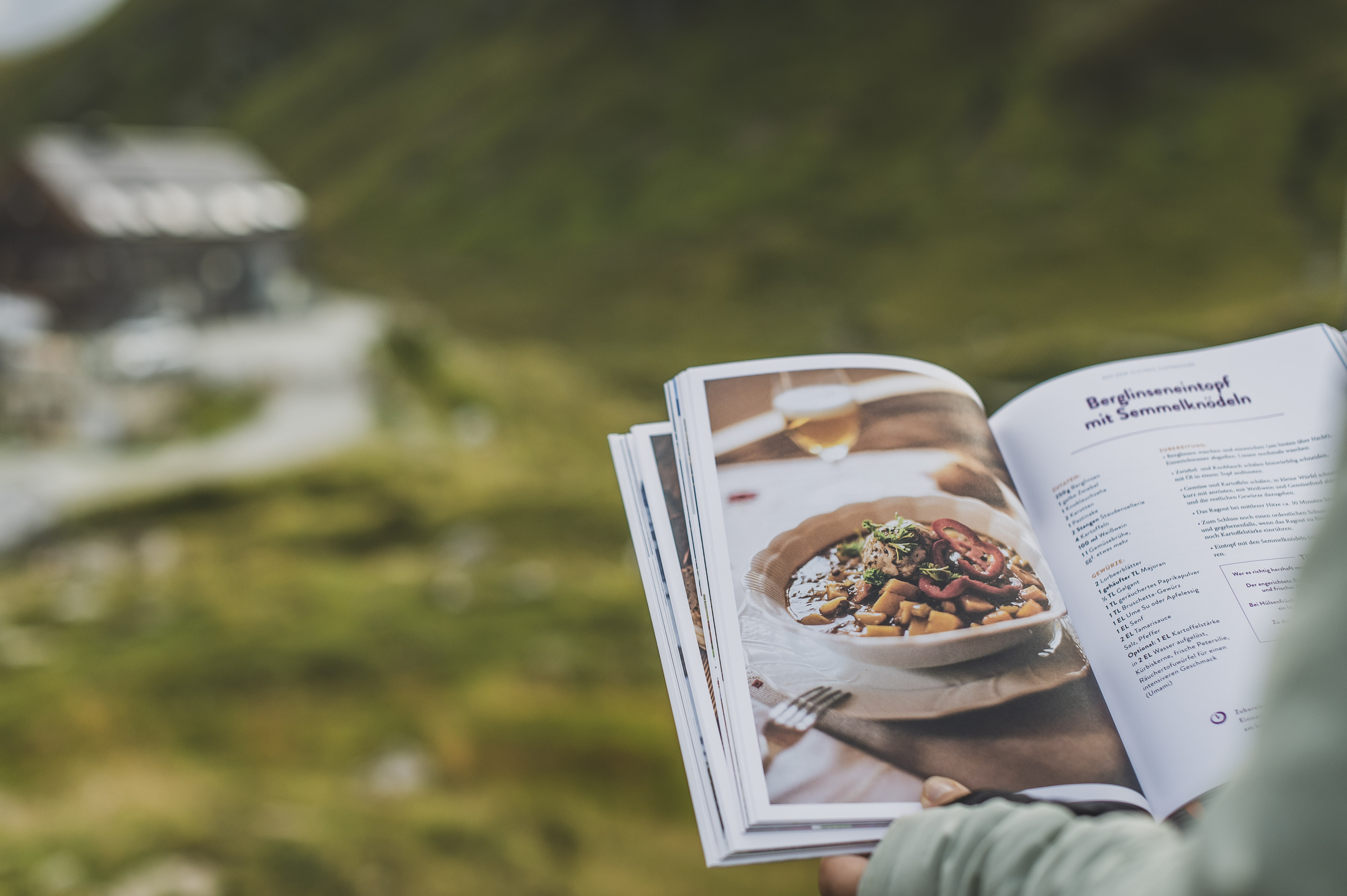 Das Kochbuch "Hüttenduft – Gerichte und Geschichten aus den Tauern" von Evelyn Matejka und Tom Burger vor der Franz-FischerHütte und Berglandschaft der Hohen Tauern.