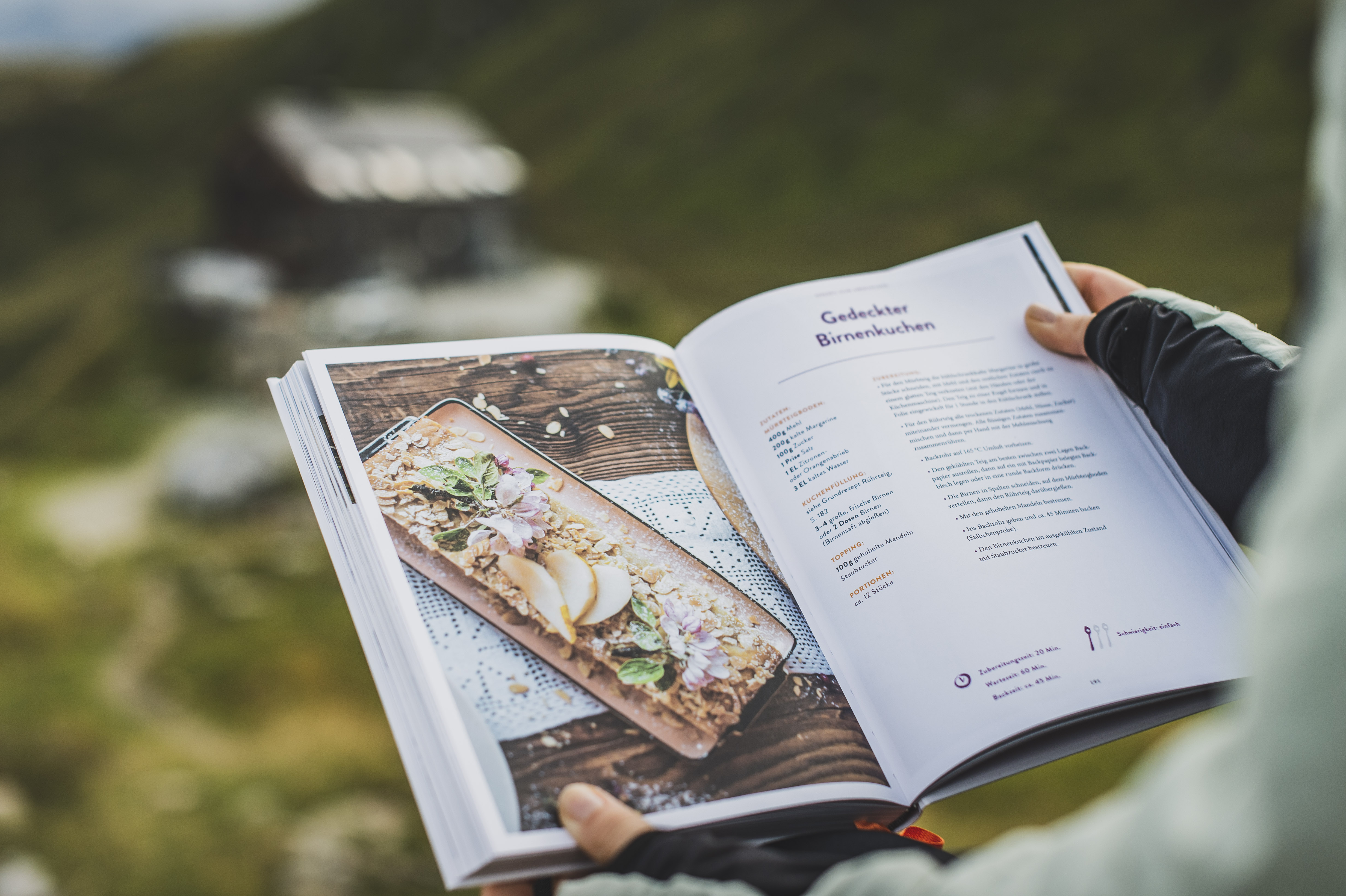 Das Kochbuch "Hüttenduft – Gerichte und Geschichten aus den Tauern" von Evelyn Matejka und Tom Burger vor der Franz-FischerHütte und Berglandschaft der Hohen Tauern.