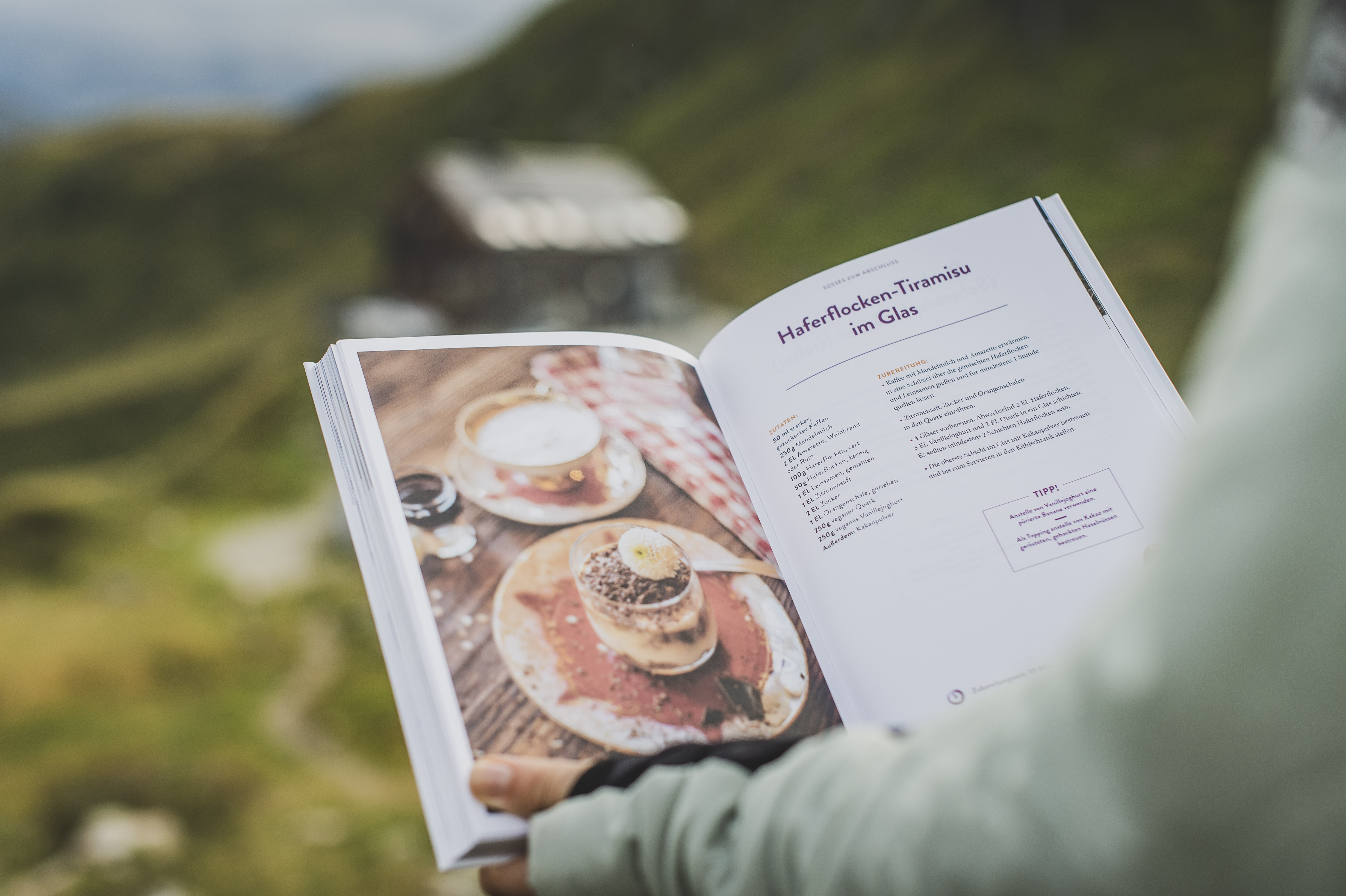 Das Kochbuch "Hüttenduft – Gerichte und Geschichten aus den Tauern" von Evelyn Matejka und Tom Burger vor der Franz-FischerHütte und Berglandschaft der Hohen Tauern.